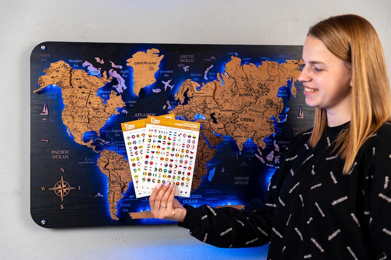 A woman is holding a map of the world with various country flags and icons on it, standing in front of a wall with a large world map.