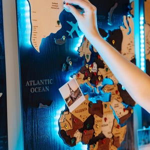 May include: A wall-mounted wooden world map with a dark blue background and integrated blue LED lighting. The map features laser-cut wooden continents and countries with text labels, decorated with colorful push pins and a small photograph of the Eiffel Tower.