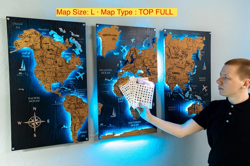 A man is holding a small set of cards in front of a wall with three large world map posters.