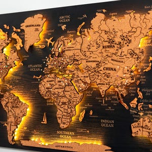 以下が含まれることがあります： 黒い背景にコルクで作られた世界地図。大陸を照らすLEDライト付き。地図には国の輪郭と海の名前が含まれています。装飾的な飛行機とヨットのアイコンが含まれています。手は地図上の場所を指しています。