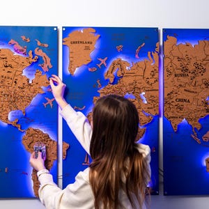 Puede incluir: Un mapa del mundo de madera de tres paneles con fondo azul y continentes marrones. El mapa tiene retroiluminación LED e incluye detalles como aviones y veleros. Una persona interactúa con el mapa.