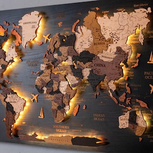 Puede incluir: Un mapa del mundo de madera multicapa para pared con varios tonos de madera marrón y negra. El mapa incluye nombres de países grabados, una rosa de los vientos e iconos decorativos de aviones y veleros, iluminado con luz LED cálida.