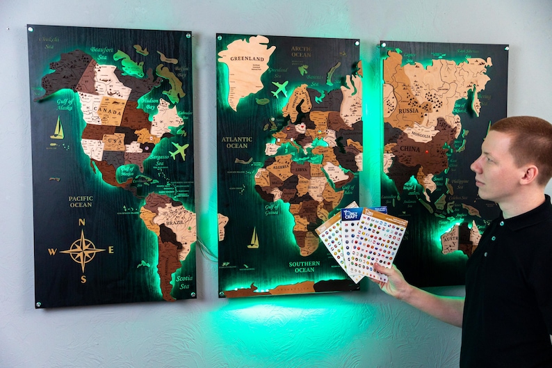 A man is standing in front of three large, illuminated world map posters on a wall, holding some stickers.