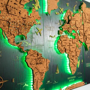 Peut inclure: Une carte du monde en bois avec un fond sombre et un rétroéclairage LED vert. La carte présente des contours de pays détaillés dans un ton de bois plus clair, avec des accents décoratifs d'avion et de voilier. Les mots "Pacific Ocean" et "Southern Ocean" sont visibles.