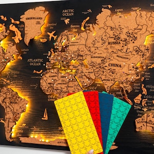 Peut inclure: Une carte du monde en bois avec une surface en liège, éclairée par des lumières chaudes. La carte est montée sur un fond noir et comprend des épingles indiquant des lieux. Des cartes de voyage colorées sont tenues devant la carte.