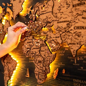 May include: A detailed cork world map with carved continents and countries, illuminated by warm lights. A hand is placing a pin on the map, marking a location. The map is mounted on a dark wooden background, creating a striking display.