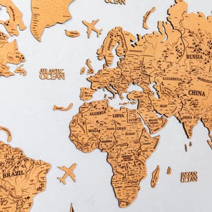 May include: A wooden world map with country outlines and labels, including "BRAZIL", "CHINA", and "RUSSIA". The map features small wooden airplane and boat cutouts. The map is a light brown color and is mounted on a white surface.