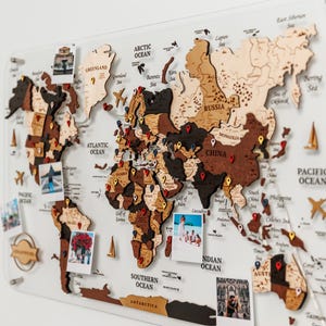 以下が含まれることがあります： 透明なアクリル板にマウントされた木製の世界地図。 地図は茶色とタンのさまざまな色合いで、場所を示すピンが付いています。 小さな写真が地図に添付されており、海にはラベルが付いています。