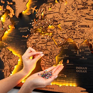 Peut inclure: Une carte du monde en bois avec un fond noir et un revêtement en liège. La carte est éclairée par des lumières blanches chaudes et comprend des punaises colorées. La carte montre les continents et les océans.