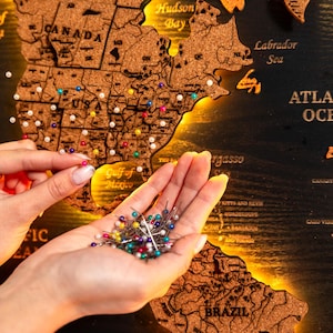 Peut inclure: Une carte du monde en liège avec des punaises, montée sur une surface en bois foncé. La carte présente des étiquettes géographiques, dont l'océan Atlantique. Une main tient une sélection de punaises colorées, prêtes à être placées sur la carte.