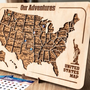 May include: A wooden map of the United States with the text "Our Adventures" and "United States Map" engraved on it. The map is divided into states and has a cork-like texture. There are small blue and green pins placed on the map.