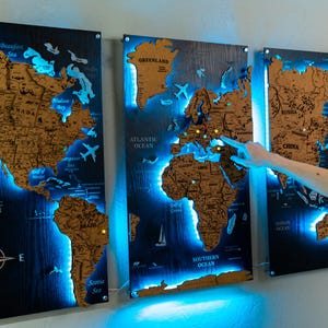 May include: Three wooden world maps with a dark blue background and illuminated blue LED lights. The maps are made of a light brown material, with the continents and oceans clearly defined. A compass rose is visible on the left map.