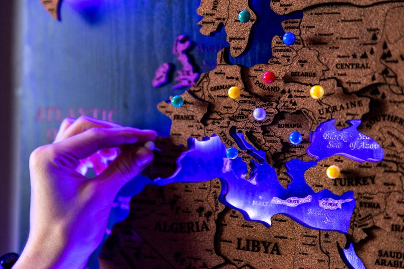 A hand is placing a blue pushpin on a cork world map, which is illuminated with blue light.