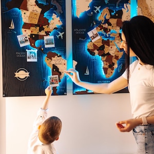 Puede incluir: Mapa del mundo de madera con retroiluminación LED. El mapa presenta contornos detallados de países y está adornado con pequeñas fotos. El mapa está montado sobre un fondo oscuro, con los océanos etiquetados. Un niño y un adulto interactúan con el mapa.