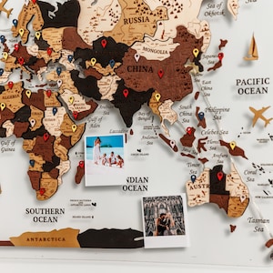 Peut inclure: Une carte du monde en bois avec des pays en nuances de marron, beige et noir. La carte comprend des marqueurs à épingles rouges, jaunes et bleus, de petites découpes d'avions et de voiliers, et plusieurs petites photographies. La carte est montée sur une surface acrylique transparente.