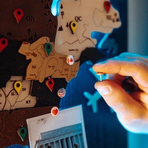 A hand is placing a pushpin on a wooden map of the United States, with various locations marked on it.