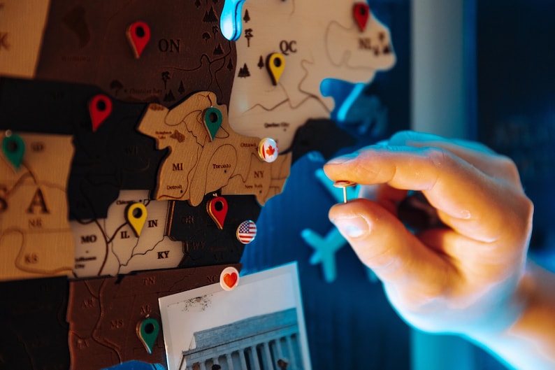A hand is placing a pushpin on a wooden map of the United States, with various locations marked on it.