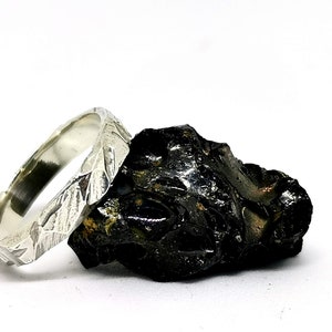 K&ouml;nnte beinhalten: Ein silberner Ring mit einer strukturierten, geh&auml;mmerten Oberfl&auml;che. Der Ring liegt auf einem dunklen, rauen Stein.