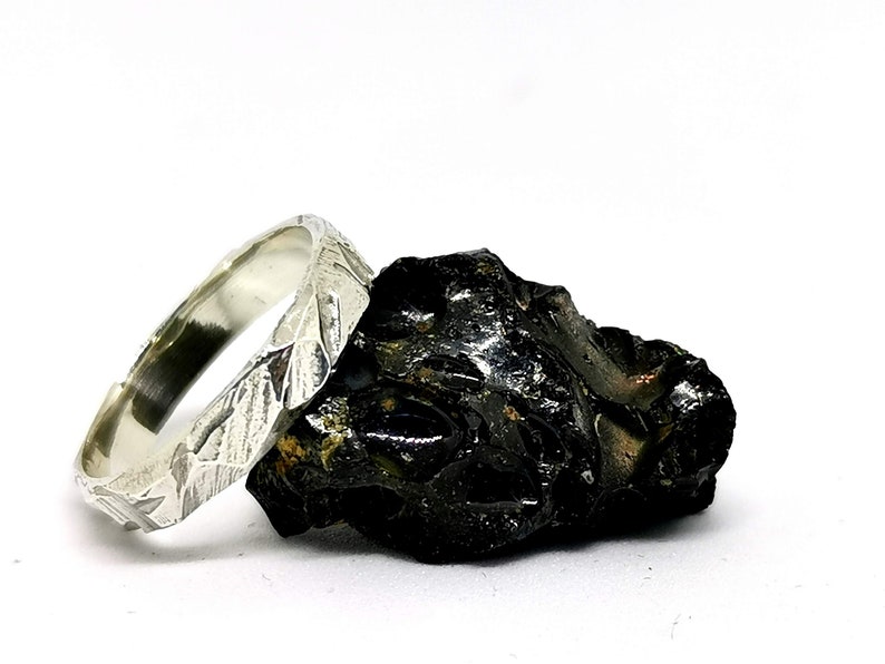K&ouml;nnte beinhalten: Ein silberner Ring mit einer strukturierten, geh&auml;mmerten Oberfl&auml;che. Der Ring liegt auf einem dunklen, rauen Stein.