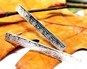 Könnte beinhalten: Ein Paar silberne Ohrringe mit Holzmaserung. Die Ohrringe sind lang und schmal und liegen auf einem braunen Blatt.