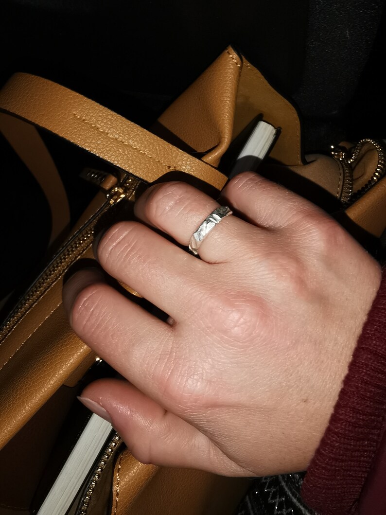 K&ouml;nnte beinhalten: Eine Hand mit einem silbernen Ring mit strukturiertem Design. Die Hand greift in eine braune Ledertasche.