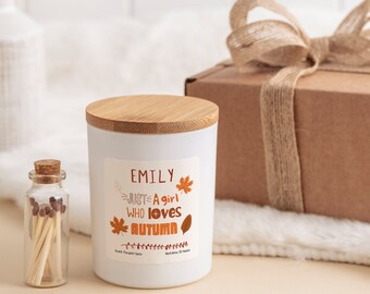 May include: A white candle with a bamboo lid and a label that reads "Emily Just A Girl Who Loves Autumn". The candle is next to a small glass jar with matches and a brown gift box with a burlap ribbon.