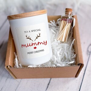 Peut inclure: Une bougie blanche avec un couvercle en bois dans une boîte en carton marron. La bougie porte une étiquette qui dit "To a special mummy Merry Christmas" avec une illustration de renne au nez rouge. Un petit bocal en verre avec des allumettes est également dans la boîte.
