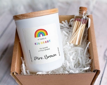 Puede incluir: Una vela blanca con una tapa de madera en una caja marrón con papel blanco triturado. La etiqueta de la vela tiene un arcoíris y el texto "Se necesita un GRAN CORAZÓN para moldear las PEQUEÑAS MENTES Sra. Brown". También hay un pequeño frasco de vidrio con cerillas en la caja.