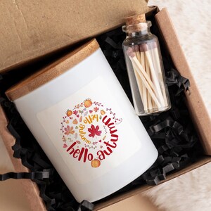 Pode incluir: Uma vela branca com tampa de madeira em uma caixa marrom com papel picado preto. A vela tem um adesivo com o texto "hello autumn" e um design de folhas de outono, abóboras e corações. Também há um pequeno frasco de vidro com fósforos na caixa.