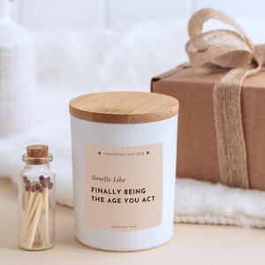 Peut inclure: Une bougie blanche dans un contenant cylindrique avec un couvercle en bambou et une étiquette beige portant l'inscription : Hand poured with love, Smells Like FINALLY BEING THE AGE YOU ACT, Vegan soy wax. Une petite bouteille en verre avec des allumettes.
