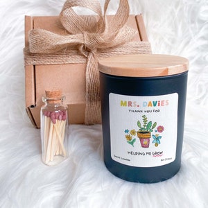 May include: A black candle with a wooden lid and a label that says "Mrs. Davies Thank You For Helping Me Grow" with a colourful illustration of a potted plant. The candle is packaged in a brown box with a hessian ribbon and a small glass jar of matches.
