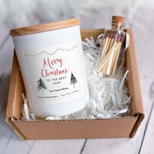 Op de afbeelding: Een witte kaars met een houten deksel en een label met de tekst "Merry Christmas to the best Mum love from Olivia" in rode en zwarte letters. De kaars zit in een bruine kartonnen doos met wit geribbeld papier. Een klein glazen potje met een kurken deksel en lucifers zit ook in de doos.