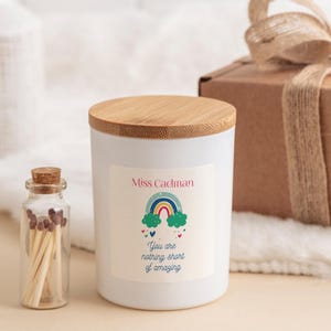 May include: White candle with a bamboo lid and a rainbow sticker that says "Miss Cadman You are nothing short of amazing". The candle is next to a small glass jar with matches.