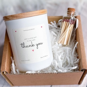 Op de afbeelding: Een witte kaars met een houten deksel en de tekst "thank you" op het etiket, gepresenteerd in een kartonnen doos met een kleine glazen fles lucifers. De kaars is geëtiketteerd met "Sweet Lavender".
