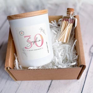 Könnte beinhalten: Eine weiße Kerze mit einem Holzdeckel in einer braunen Box mit weißem, zerkleinertem Papier. Die Kerze hat ein Etikett mit der Nummer 30 und dem Namen Sophia, mit den Worten "Happy Birthday". Ein kleines Glas mit Streichhölzern befindet sich ebenfalls in der Box.