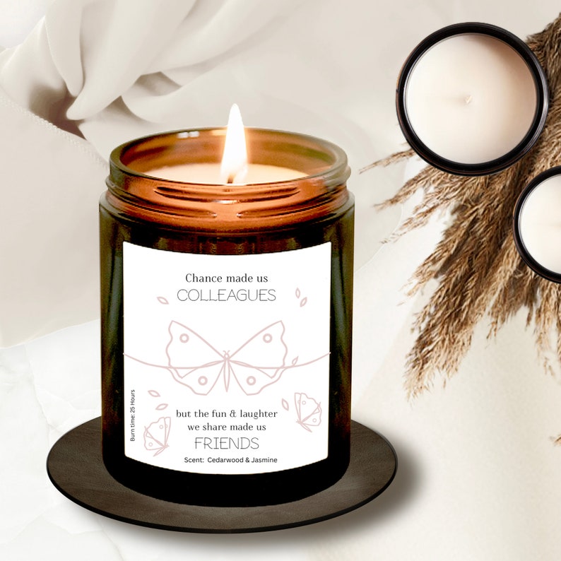 May include: A brown glass candle with a white label that reads "Chance made us colleagues but the fun & laughter we share made us friends". The label also features a butterfly design and the text "Cedarwood & Jasmine" for the scent.