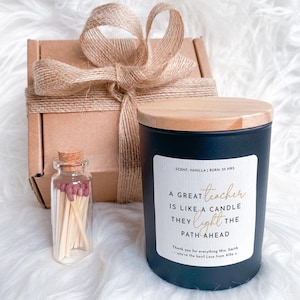 May include: A gift set featuring a black vanilla-scented candle with a wooden lid, a small glass bottle of matches, and a wrapped cardboard box. The candle has a label with gold text that reads, "A great teacher is like a candle they light the path ahead."