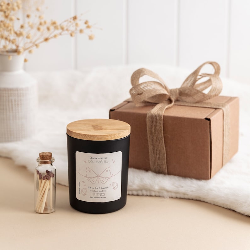 May include: A black candle with a wooden lid and a white label that reads "Chance made us colleagues, but the fun & laughter we share made us friends." The candle is sitting on a white fluffy surface next to a small glass jar with matches and a brown gift box with a hessian ribbon.