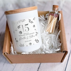 Op de afbeelding: Een witte kaars met een houten deksel en een label met de tekst "15 Crystal Wedding Anniversary Martha & Arthur 23.05.2010". Een klein glazen flesje gevuld met lucifers staat naast de kaars in een kartonnen doos gevuld met witte papiersnippers.