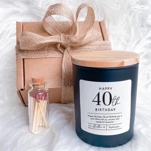 Könnte beinhalten: Eine schwarze Kerze mit Holzdeckel und einem weißen Etikett mit der Aufschrift "HAPPY 40th BIRTHDAY". Eine kleine Glasflasche mit Streichhölzern und eine verpackte Geschenkbox sind ebenfalls vorhanden. Die Kerze ist Teil eines Geschenksets.
