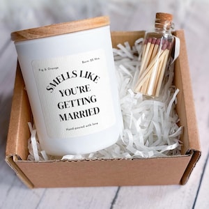May include: A white candle with a wooden lid and a label that reads "Smells Like You're Getting Married" sits in a cardboard box with a small glass bottle of matches. The candle is labeled "Fig & Orange" and "Burn: 25 Hrs."