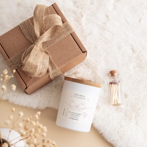 May include: A white candle with a wooden lid and a pink butterfly design. The candle has the text "Dance made us colleagues, but the fun & laughter we share made us friends" on the label. The candle is packaged in a brown box with a brown ribbon and a small glass jar with matches.