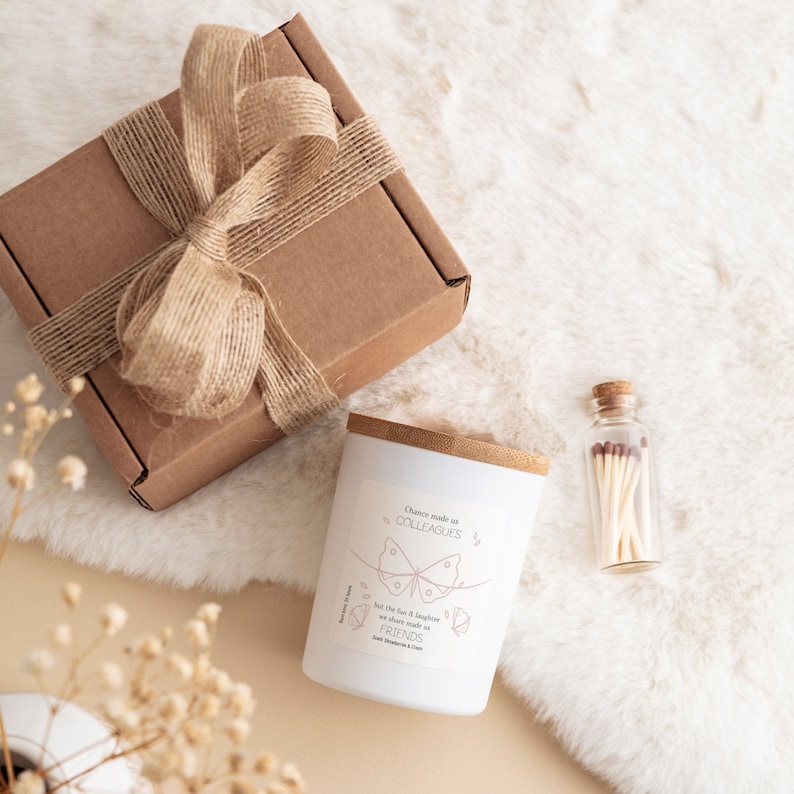 May include: A white candle with a wooden lid and a pink butterfly design. The candle has the text "Dance made us colleagues, but the fun & laughter we share made us friends" on the label. The candle is packaged in a brown box with a brown ribbon and a small glass jar with matches.