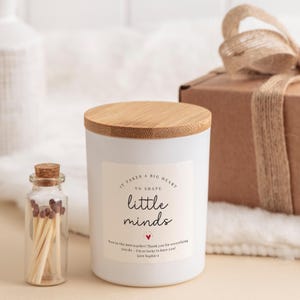 May include: A white candle with a wooden lid and the text "It takes a big heart to shape little minds." A small glass bottle of matches is next to the candle. A wrapped gift box is in the background.
