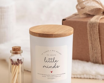 Puede incluir: Una vela blanca con tapa de madera y el texto "It takes a big heart to shape little minds". Una pequeña botella de vidrio con cerillas está al lado de la vela. Una caja de regalo envuelta está en el fondo.