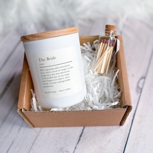 May include: A white candle with a wooden lid and a label that reads "The Bride" with a description about the bride's excitement and a new adventure. The candle is in a brown box with white shredded paper and a small glass jar with matches.