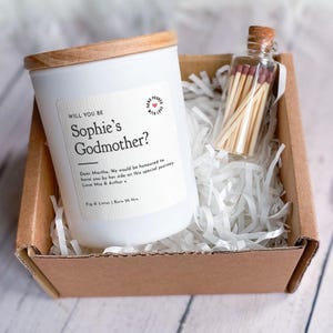 以下が含まれることがあります： 「Will You Be Sophie's Godmother?」と書かれたラベルが付いた、木製の蓋が付いた白いキャンドルが、白い紙の詰め物が入った段ボール箱に入っています。マッチの小さなガラス瓶も含まれています。