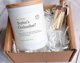 May include: A white candle with a wooden lid and a label that reads "Will You Be Sophie's Godmother?" is presented in a cardboard box with white paper filler. A small glass bottle of matches is also included.