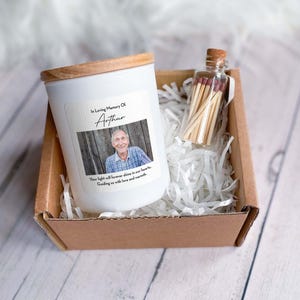 May include: A white candle with a wooden lid in a brown box with white shredded paper. The candle has a label that says "In Loving Memory Of Arthur" and includes a photo of a man. The box also contains a small glass jar with matches.
