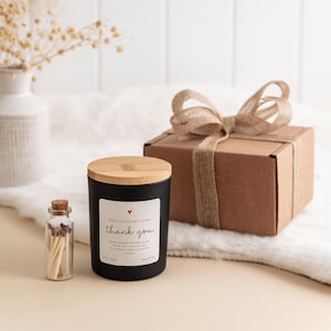 Peut inclure: Une bougie noire avec un couvercle en bois et une &eacute;tiquette &laquo;&nbsp;merci&nbsp;&raquo;, &agrave; c&ocirc;t&eacute; d'une petite bouteille en verre d'allumettes et d'un coffret cadeau emball&eacute; avec un n&oelig;ud en toile de jute. La bougie est sur une surface blanche et moelleuse.